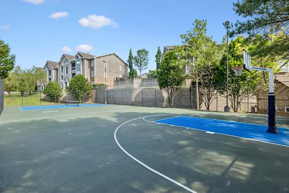 A basketball court with a blue surface and white lines, surrounded by a fence and houses.