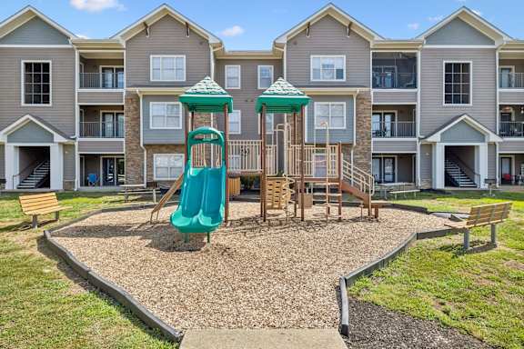 A playground with a green slide and wooden structure in front of apartment buildings.
