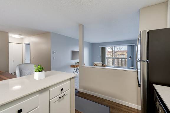 A kitchen with a refrigerator, white countertop, and white cabinets with a view over a half wall into a living room.
