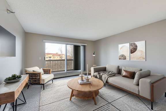 A living room with a sofa, chairs, and a coffee table, and access to the balcony via sliding glass door.