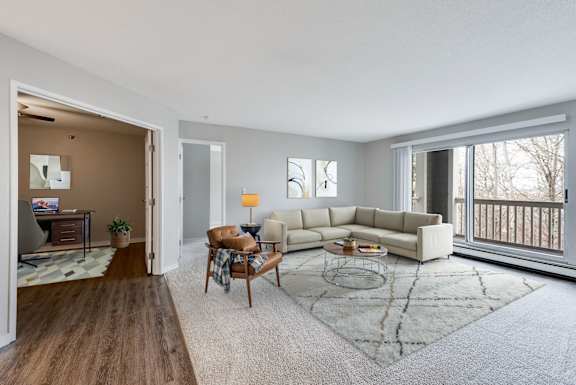 A large, carpeted living room with a sliding glass door onto a balcony and a view into a study with a desk.