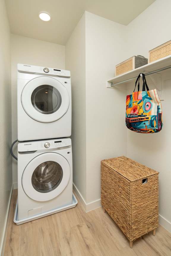 A small laundry room with a double washer and dryer, a wicker basket and a bag hanging on the wall.
