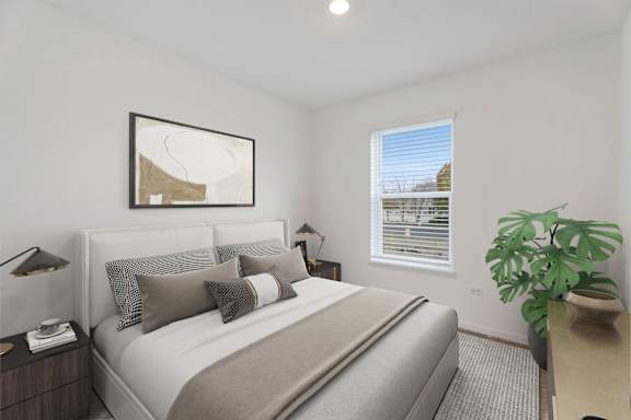 A bedroom with a large bed, a nightstand, a lamp, a plant, and a framed picture on the wall.