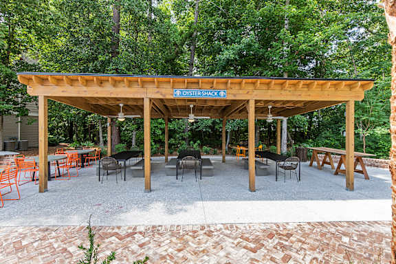 A wooden pavilion with tables and chairs is surrounded by trees.