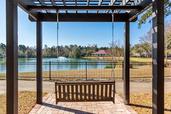 A wooden bench is under a pergola with a view of a pool and trees.