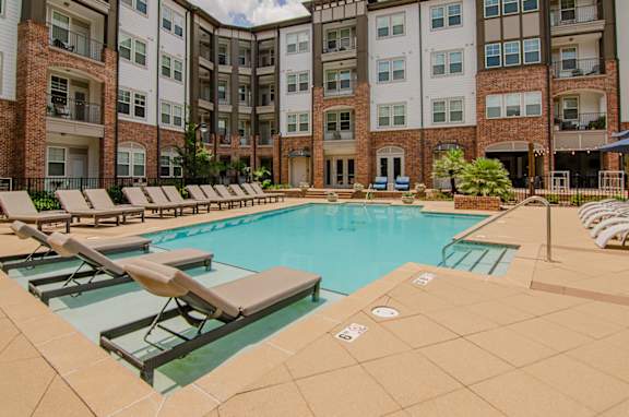 A large swimming pool surrounded by lounge chairs in front of apartment buildings.