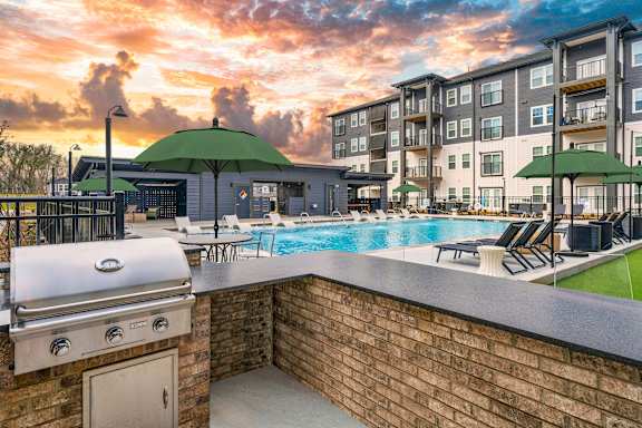 A sunset view of a pool area with a barbecue grill and lounge chairs.