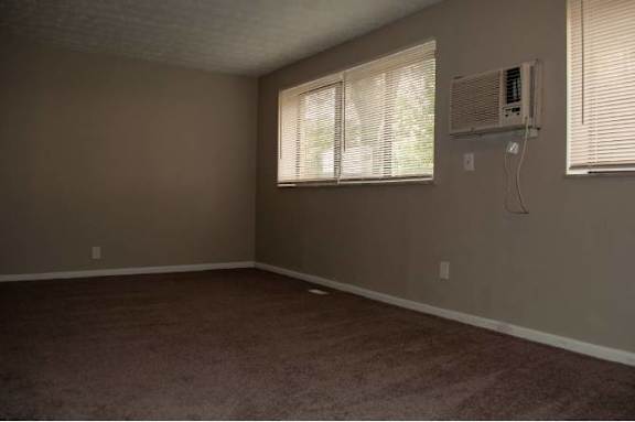 A room with a carpeted floor, a window with blinds, and a wall-mounted air conditioning unit.