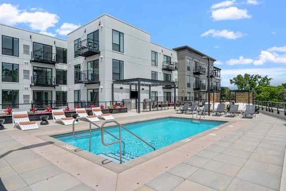 A swimming pool is surrounded by lounge chairs in front of apartment buildings.