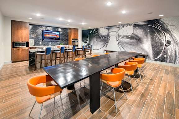 A black and white photo of a woman's face is on the wall behind a long black table with orange chairs.