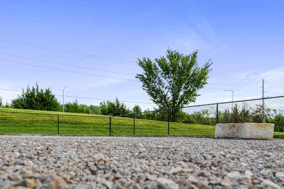 A gravel area with a fence, a tree, and a stone in the foreground.