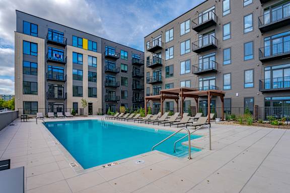 A swimming pool is surrounded by lounge chairs in front of apartment buildings.