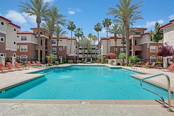 A resort-style swimming pool here at Mozaic at Steele Park surrounded by tall palm trees, comfortable lounge chairs, and shaded patio seating. The sparkling blue water is framed by spacious sundecks and modern apartment buildings, creating a relaxing outdoor retreat perfect for soaking up the Arizona sunshine or enjoying time with neighbors.