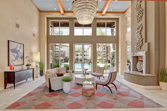 A grand clubhouse lounge here at Mozaic at Steele Park featuring soaring ceilings with exposed wood beams, a sparkling chandelier, and expansive windows that fill the space with natural light. A stylish seating arrangement rests on a patterned area rug near a modern fireplace, creating an elegant and welcoming atmosphere with views of the pool courtyard just beyond the glass doors.