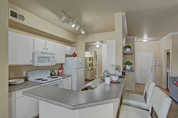A spacious kitchen here at Mozaic at Steele Park featuring white cabinetry, generous counter space, and a breakfast bar with seating. Matching white appliances including a built-in microwave, range, and refrigerator create a cohesive look, while track lighting and the open layout connect seamlessly to the adjacent living area.