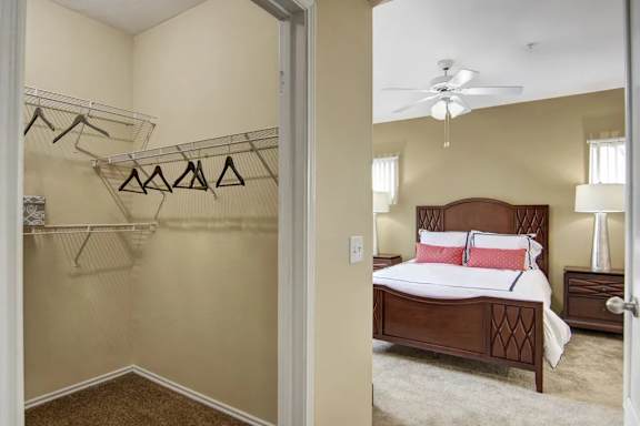 A spacious bedroom with walk-in closet here at Mozaic at Steele Park featuring plush carpet, neutral walls, and a ceiling fan with light. The open closet includes built-in shelving and hanging space, while the adjoining bedroom comfortably fits a full bedroom set with matching nightstands for added storage.