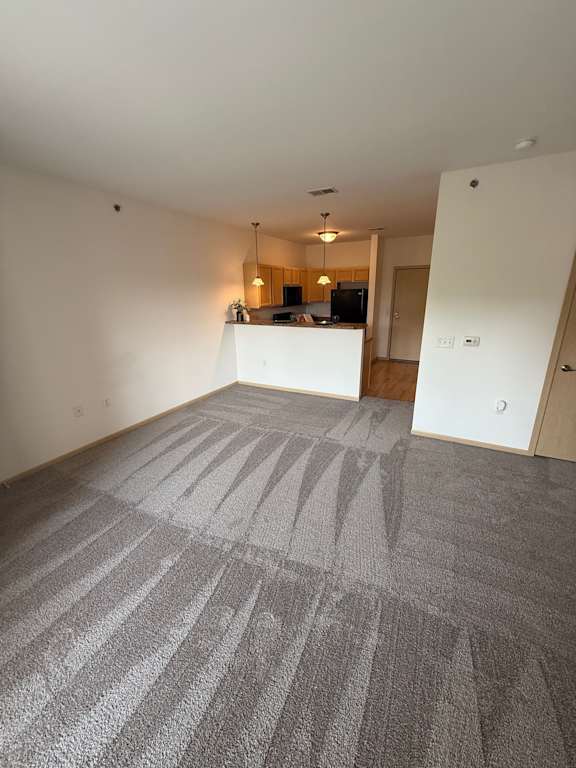 A room with a carpeted floor and a kitchen area in the background.