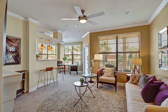 A bright, open living room here at Tuscany at Faudree with plush carpeting, a comfortable sofa, and accent chairs arranged around a patterned area rug. Large windows with blinds bring in natural light, while a ceiling fan, table lamps, and neutral décor create a warm, inviting atmosphere that flows into a nearby dining area.