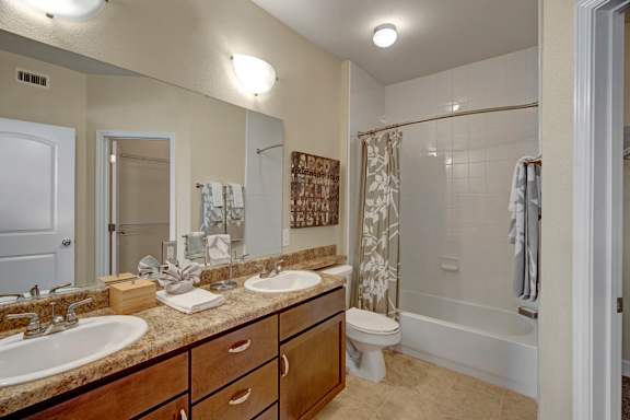 A clean, spacious bathroom here at Tuscany at Faudree with a long vanity featuring a light countertop, double sinks, and wood cabinetry. A large wall mirror and overhead lighting brighten the space, while a bathtub and shower combo with tiled walls sits beside a toilet. Neutral finishes and tiled flooring create a fresh, comfortable bathroom layout.