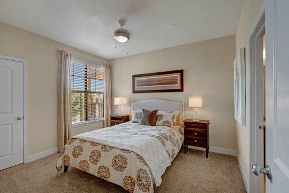 A spacious bedroom here at Tuscany at Faudree with plush carpet, soft neutral walls, and a comfortable bed centered between matching nightstands and lamps. A large window with blinds and curtains brings in natural light, while framed wall art and a ceiling fan add a calm, inviting feel to the room.