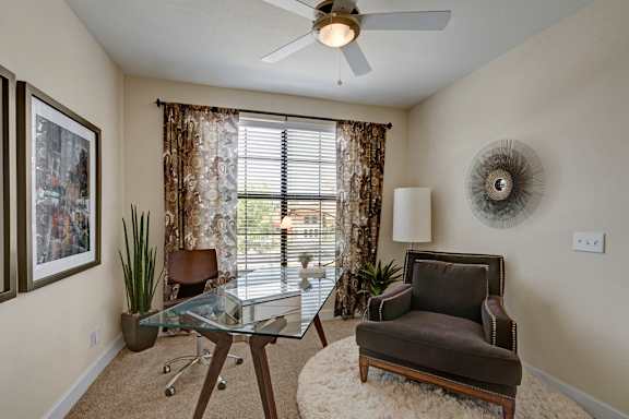 A bright home office here at Tuscany at Faudree with plush carpet, neutral walls, and a glass-top desk paired with a rolling chair. A comfortable accent chair sits on a soft area rug, while a ceiling fan and large window with blinds and patterned curtains bring in natural light and create a calm, functional workspace.