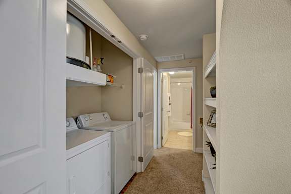A convenient in-home laundry area here at Tuscany at Faudree with a full-size washer and dryer tucked into a hallway closet. Open shelving above provides storage for supplies, while plush carpet, neutral walls, and nearby built-in display shelves create a clean, functional transition space leading toward the bathroom.