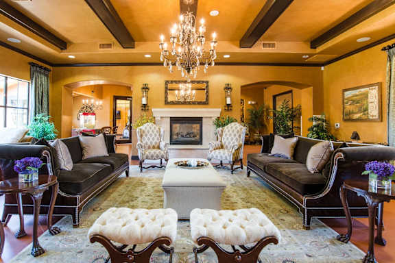 A grand community lounge here at Tuscany at Faudree with elegant seating arranged around a central ottoman and decorative rug. Plush sofas, accent chairs, and side tables create a welcoming gathering space, while a statement chandelier, fireplace, framed artwork, and lush greenery add warmth and upscale character.