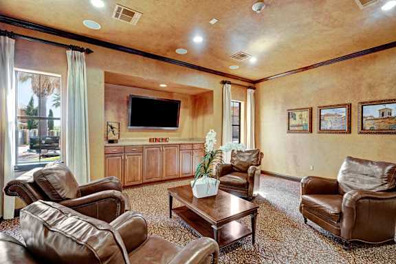 A cozy media lounge here at Tuscany at Faudree with plush leather seating arranged around a wooden coffee table. A large mounted television sits above built-in cabinetry, creating a dedicated entertainment area. Soft carpeting, framed wall art, and tall windows with curtains add warmth and natural light to the inviting community space.