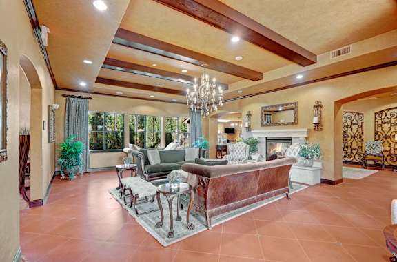 A spacious community lounge here at Tuscany at Faudree with elegant seating arranged on an area rug beneath exposed ceiling beams and a statement chandelier. Plush sofas, accent chairs, and side tables surround a decorative fireplace, while large windows, greenery, and warm finishes create an inviting space for relaxing and gathering.