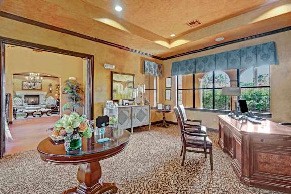A refined business center here at Tuscany at Faudree with a large wooden desk, guest chairs, and a computer workstation. Decorative tables, framed artwork, and soft carpeting add an upscale feel, while large windows bring in natural light and connect the space to adjacent community lounges.