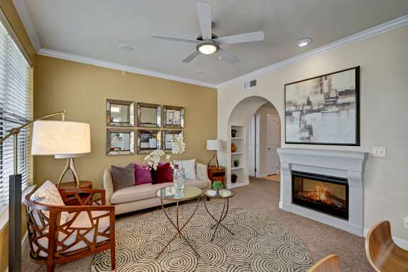A stylish living room here at Tuscany at Faudree with plush carpet, a comfortable sofa accented by pillows, and a round patterned area rug. A built-in fireplace creates a cozy focal point beneath wall art, while a ceiling fan, table lamps, and arched doorway add warmth, light, and an inviting, open feel.
