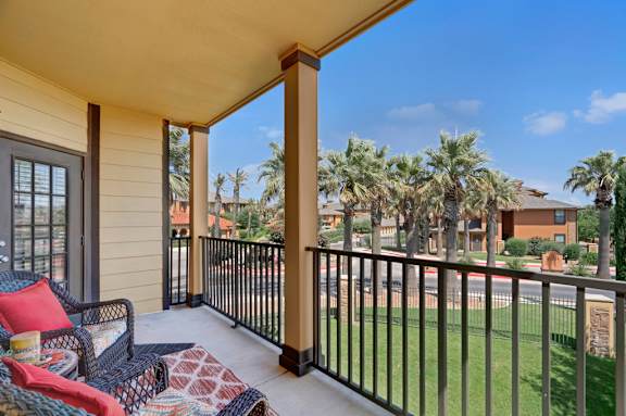 A covered private balcony here at Tuscany at Faudree with outdoor seating arranged on a concrete floor. A railing overlooks landscaped grounds with palm trees and nearby community buildings, while the shaded layout offers a comfortable space to relax and enjoy fresh air and neighborhood views.