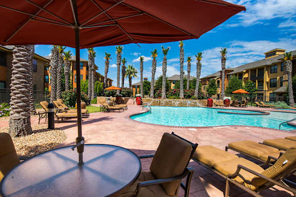 A resort-style outdoor pool area here at Tuscany at Faudree featuring a sparkling curved swimming pool surrounded by palm trees, tan lounge chairs, and shaded seating under large umbrellas. Warm-toned buildings, landscaped greenery, and a sunny blue sky create a relaxing, vacation-inspired community atmosphere.