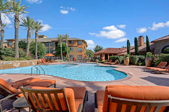 A sunny resort-style swimming pool here at Tuscany at Faudree showcasing a curved pool with clear blue water, surrounding tan pool deck, and orange cushioned lounge chairs in the foreground. Palm trees, landscaped planters, and Mediterranean-inspired buildings frame the pool area beneath a bright blue sky with scattered clouds.