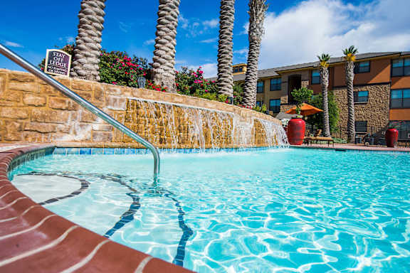 A refreshing resort-style swimming pool here at Tuscany at Faudree featuring clear blue water, a shallow entry with handrail, and a stone waterfall feature. Tall palm trees, colorful landscaping, and Mediterranean-inspired apartment buildings surround the pool beneath a bright blue sky.