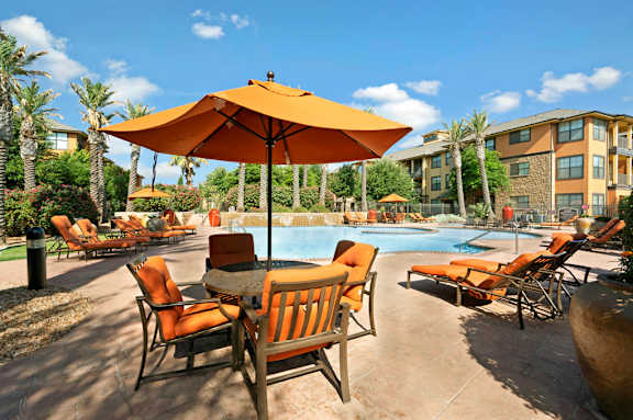 A sunny poolside gathering area here at Tuscany at Faudree featuring a resort-style swimming pool surrounded by orange cushioned lounge chairs, shaded seating with umbrellas, and a spacious tan pool deck. Palm trees, landscaped greenery, and Mediterranean-inspired apartment buildings frame the scene under a bright blue sky.