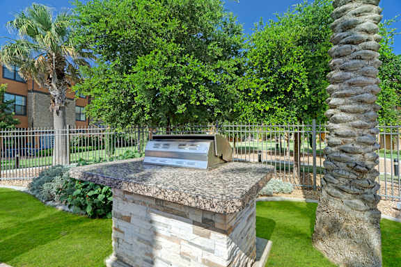 A convenient outdoor grilling area here at Tuscany at Faudree featuring a built-in stainless steel grill set on a stone base, surrounded by green lawn, palm trees, and landscaped greenery. A metal fence and nearby apartment buildings create a relaxed community setting under a clear blue sky.