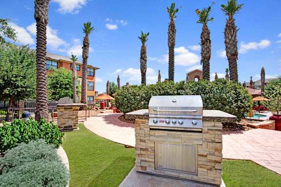 A landscaped outdoor grilling station here at Tuscany at Faudree featuring a built-in stainless steel grill on a stone base, surrounded by manicured green lawn, palm trees, and curved walkways. Mediterranean-inspired apartment buildings, poolside seating, and bright blue skies create a welcoming community atmosphere.