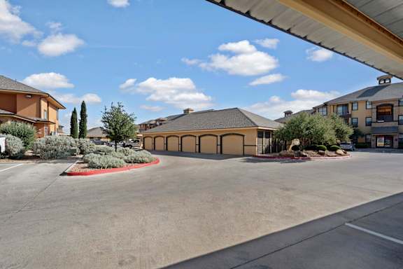 A spacious parking area here at Tuscany at Faudree featuring detached garage buildings with individual doors, wide paved drive lanes, and neatly landscaped islands with shrubs and small trees. Mediterranean-inspired apartment buildings surround the garages, creating a clean and well-organized community setting under a bright blue sky.