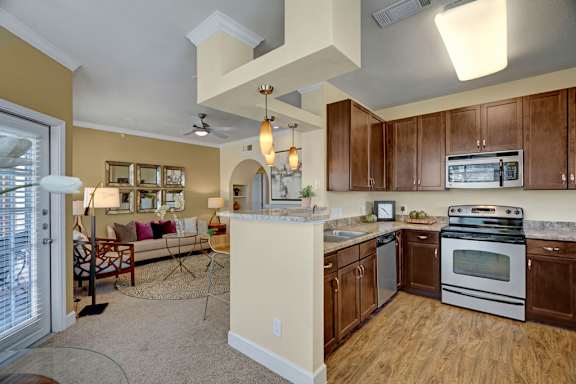 A bright open-concept kitchen and living area here at Tuscany at Faudree with rich wood cabinets, light countertops, and a full appliance setup including a stove, microwave, sink, and dishwasher. Pendant lighting highlights a breakfast bar, while plush carpet and coordinated décor create a seamless flow into the living space.