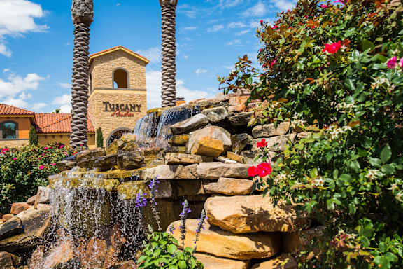 A scenic stone waterfall feature here at Tuscany at Faudree framed by lush greenery, blooming red roses, and textured rock tiers. Flowing water cascades over natural stone beneath tall palm trees, with Mediterranean-style buildings and a blue sky backdrop creating a serene, resort-inspired community entrance.