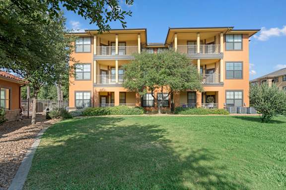 A well-maintained apartment building exterior here at Tuscany at Faudree featuring a three-story design with covered balconies, warm stucco tones, and large windows. Manicured green lawn, mature shade trees, and neatly landscaped walkways create a peaceful residential setting under a bright blue sky.