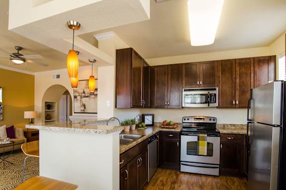 A modern open kitchen here at Tuscany at Faudree with dark wood cabinets, light granite-style countertops, and wood-look flooring. A breakfast bar with pendant lighting overlooks the living area, while a stainless steel refrigerator, stove, microwave, dishwasher, and double-basin sink create a functional and inviting cooking space.