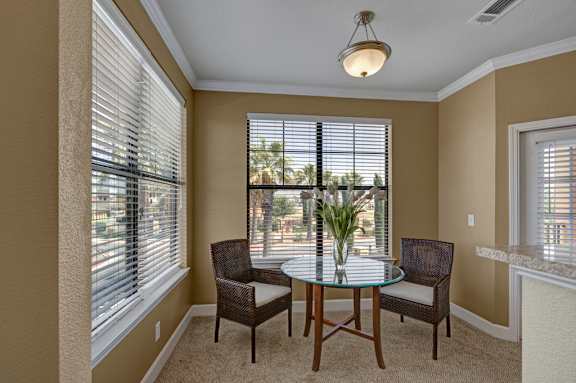 A bright dining nook here at Tuscany at Faudree with plush carpet and neutral walls. A small round glass-top table with two chairs sits beneath a ceiling light, while large windows with blinds bring in natural light and views of surrounding greenery, creating a cozy space for everyday meals.