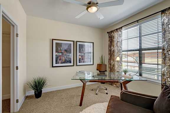 A bright home office here at Tuscany at Faudree with plush carpet, neutral walls, and a glass-top desk paired with a modern chair. Large windows with blinds and patterned curtains bring in natural light, while framed artwork, a ceiling fan, and greenery create a comfortable and functional workspace.