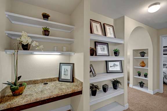 A built-in workspace and display area here at Tuscany at Faudree featuring a granite-style countertop with open shelving above and accent lighting. Nearby recessed wall shelves showcase framed artwork and greenery, while an arched hallway and plush carpet create a functional transition space with added storage and visual interest.