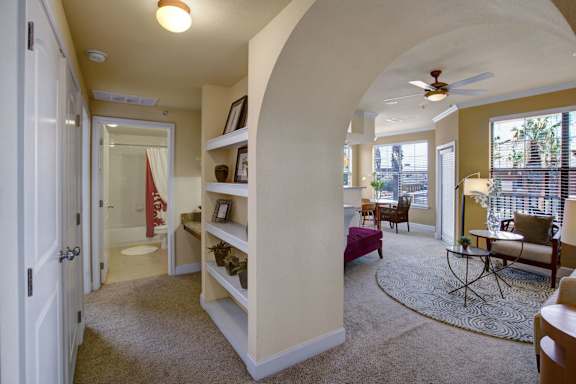 A welcoming interior hallway here at Tuscany at Faudree with plush carpet, soft neutral walls, and an arched entryway opening into the living area. Built-in display shelves add character and storage, while the layout offers clear access to the bathroom and main living space filled with natural light.