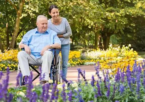 A man in a blue shirt is sitting in a wheelchair in a garden with a woman standing behind him.