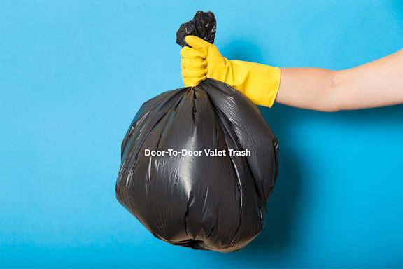 A hand in yellow rubber gloves holds a black trash bag labeled "Door-To-Door Valet Trash".