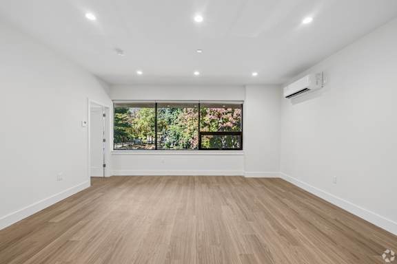 A spacious room with wooden flooring and a large window with a view of trees.